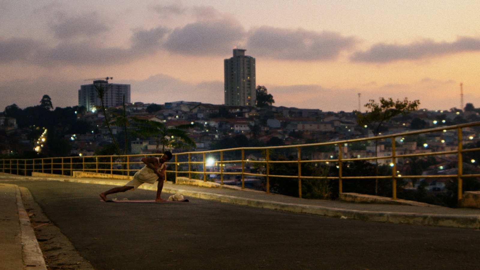 Cena do filme Yoga na Quebrada: praticante em postura de yoga numa rua em mirante, com a comunidade e o céu ao entardecer ao fundo