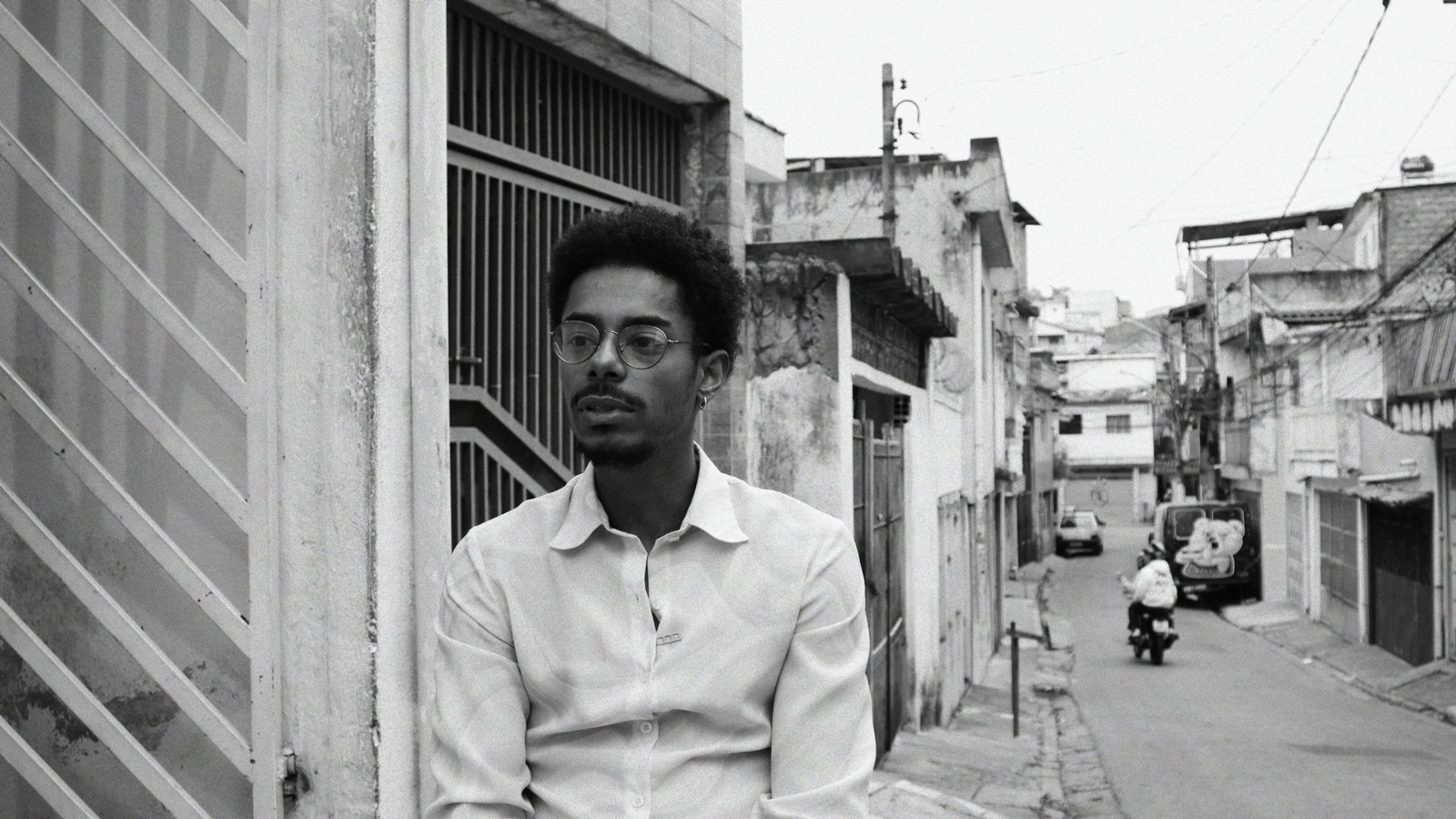 Cena em preto e branco do filme: retrato do protagonista de óculos numa viela da comunidade